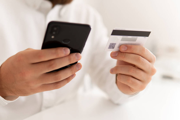 Online πληρωμή, Closeup του νεαρού άνδρα που κάνει Πληρωμή online ψώνια smartphone στο χώρο εργασίας. Αγορές ιστού. Ανδρικά χέρια κρατώντας μια πιστωτική κάρτα Man χέρι πιστωτική κάρτα για ψώνια σε απευθείας σύνδεση. - Φωτογραφία, εικόνα