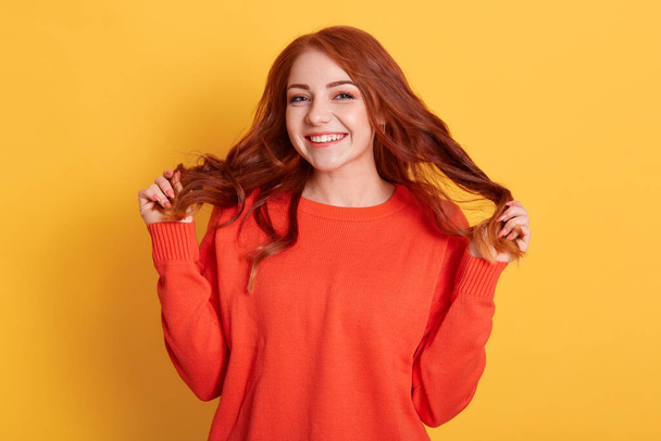 Good looking redhead happy woman smiling, touching her hair and gazing to camera with delighted expression, laughing and standing against yellow background in orange sweater. - Фото, зображення