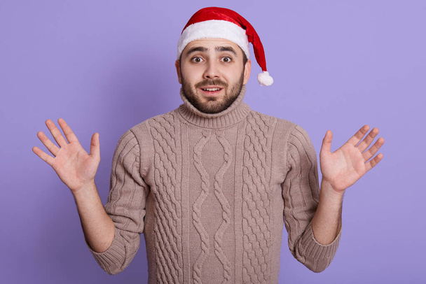 Astonished bearded man wearing Santa hat and beige pullover standing with opened mouth and big eyes, looking at camera over lilac background, raising palms up. - Fotografie, Obrázek