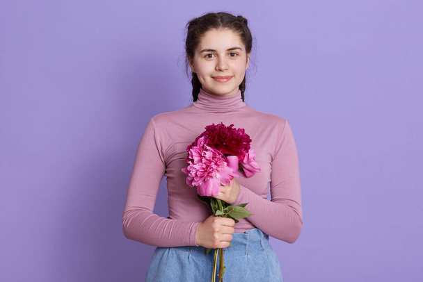 Cute smiling nice young dark haired girl in casual shirt and jeans, female with pigtails holding bouquet of peony flowers, looks at camera, has pleasant appearance, being in good mood. - Photo, image