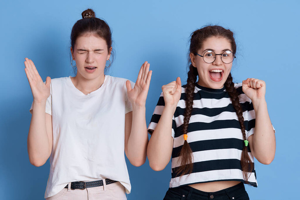 Photo of two emotional girls friends in casual t shirts, excited teenage girl wearing eyeglasses clenches fists and exclaims, female with knot keeps eyes closed, raising palms, looks displeased. - Foto, Bild