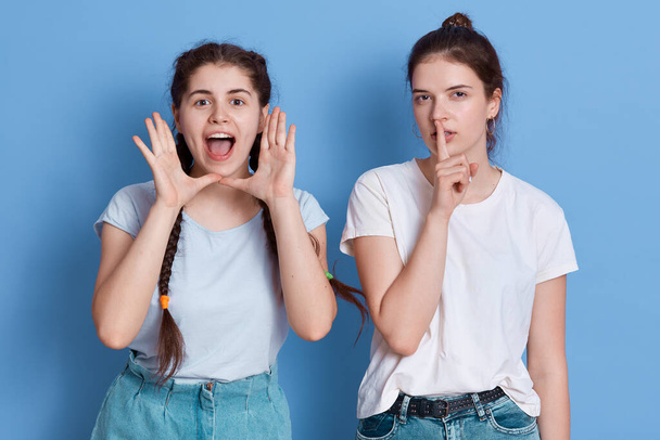 Brunette lady with hair bun refuses to listen to cry of her friend and keeps finger near lips, female with pigtails exclaiming happily and loudly, isolated over blue background. - Foto, imagen