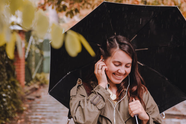 jeune jolie femme portrait écouter de la musique avec casque tout en marchant dans la rue avec parapluie sous la pluie. espace de copie - Photo, image
