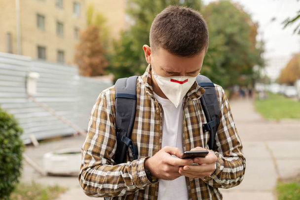 Young guy in a protective mask uses the phone outdoors - Photo, Image