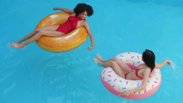 Two persons exotic mixed race girls lie sit in colored inflatable swimming rings, whirl in water, swim in pool in camp hotel resort, laugh, smile, hold hands, enjoy time together, friendship concept - Footage, Video