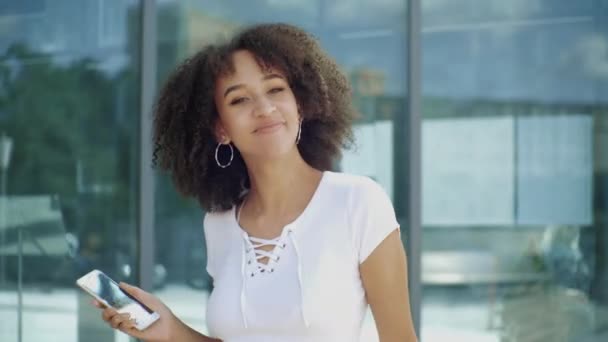 Happy joyful young afro american millennial woman enjoys jumping high, waving arms and dancing in air with smartphone in hands, laughing smiling at fun against backdrop of modern urban city building. - Footage, Video
