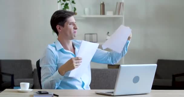 Happy male office worker manager clerk sits at office table, holds documents in his hands, joyfully actively carelessly scatters sheets of paper orders agreement in air, rejoices playing at workplace - Footage, Video