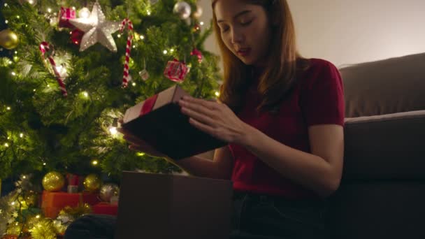 若いアジアの女性は自宅のリビングルームで装飾されたクリスマスツリーの近くにX'Masプレゼントボックスを開いて楽しんでいます。メリークリスマスの夜とハッピーニューイヤーの休日の祭り. - 映像、動画