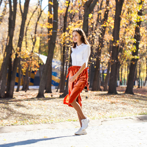 Full body portrait of a young brunette woman in orange skirt walking in autumn park - Photo, Image