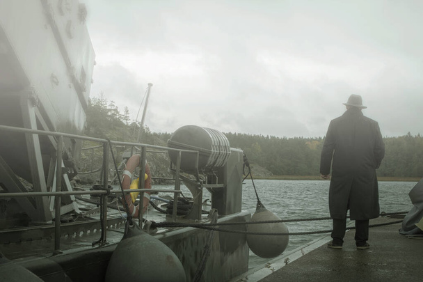 Nierozpoznawalny człowiek w kapeluszu w deszczu patrzący jak mgła wypływa z morza, noir czuje. - Zdjęcie, obraz