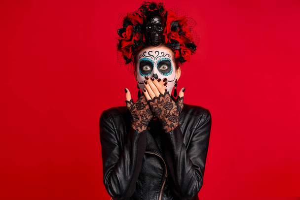 Girl with sugar skull makeup with a wreath of flowers on her head and skull, wearth black gloves close her opened shocked mouth isolated on red background. concept of Halloween or La Calavera Catrina. - Foto, Imagen