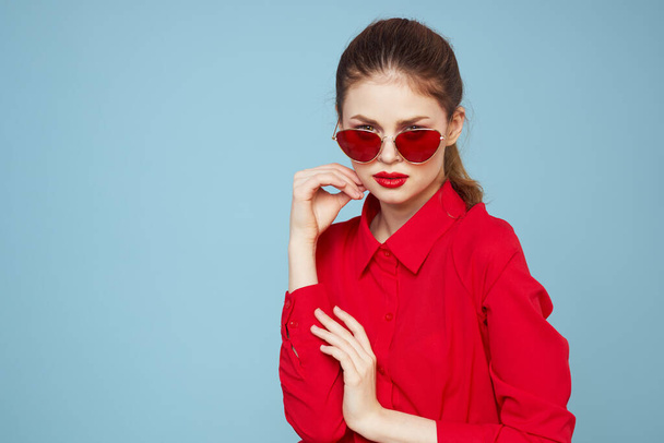 Woman in red shirt on blue background holds hands near face and sunglasses red lips makeup model - Foto, Imagen
