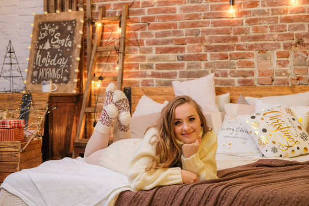 Young, smiling woman on bed at home. Loft style christmas cozy decor on the background. Merry Christmas and Happy Holidays. Blonde, cute, woman in white sweater and warm scandinavian socks. New Year's Eve mood. - Fotoğraf, Görsel