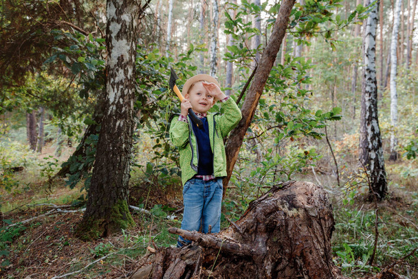 Pikkupoika hatussa syksyn metsässä. Kirveellinen poika hakkaa puuta. Pikku metsuri. Työskentele metsässä. - Valokuva, kuva