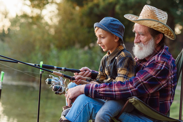 Likable respected 70-aged bearded grandfather holding on knees 10-aged cute grandson and teaching him to catch fish. - Photo, image