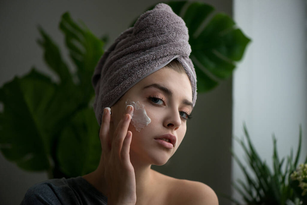 Young woman applying face cream on her face. Beauty model with perfect fresh skin and long eyelashes cares about her skin at home. Spa and Wellness, Skin Care Concept. Close up, selected focus. - Фото, изображение