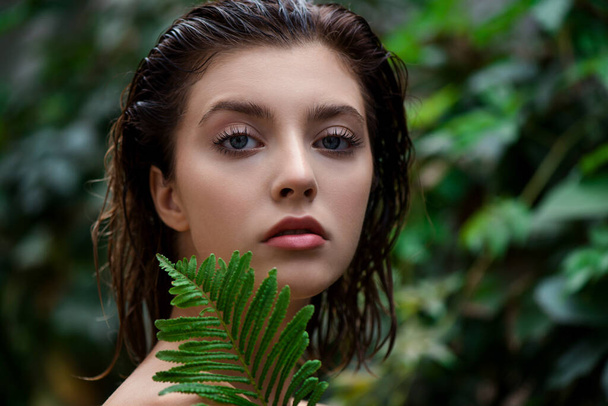Beautiful young woman with perfect skin and natural make up posing front of plant tropical green leaves background with fern. Teen model with wet hair care of her face and body. SPA, wellness, bodycare and skincare.  Close up, selective focus. - Foto, imagen
