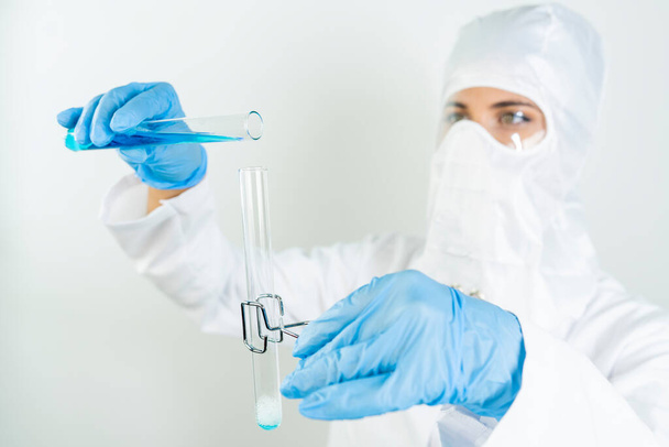 Молода концентрована жінка-вчена в захисних окулярах, масці і рукавичках скидає синю рідку речовину в пробірку з піпеткою в науковій хімічній лабораторії
    - Фото, зображення