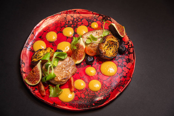 Warm salad, fried pork, fig slices, fried peaches, blueberries, cherry tomato slices, green leaves, pumpkin puree sauce. On a handmade red plate and a dark background. - Photo, Image
