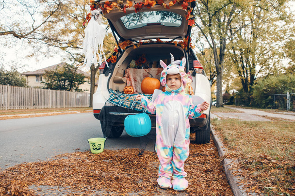 Trick or trunk. Sad upset baby in unicorn costume celebrating Halloween in trunk of car. Cute toddler preparing for October holiday outdoor. Social distance and safe alternative celebration. - Photo, Image
