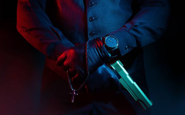 Foto eines männlichen Mafia-Verbrechers in Anzug und Lederhandschuhen, der eine Pistole mit Kreuz auf schwarzem Hintergrund hält. - Foto, Bild