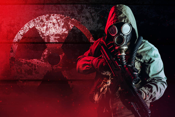 Foto de un soldado acosador apocalíptico en máscara de gas y chaqueta de campana sosteniendo rifle y de pie en la pared de metal oxidado oscuro con signo de radiación y luz brillante roja. - Foto, imagen