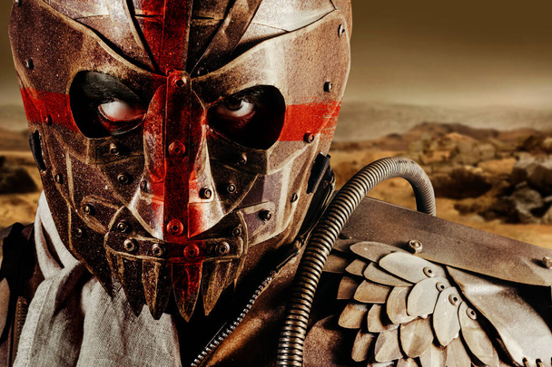 Photo of a post apocalyptic raider warrior in leather jacket with metal armor and steel mask with red cross painting standing in desert wasteland. - Foto, imagen