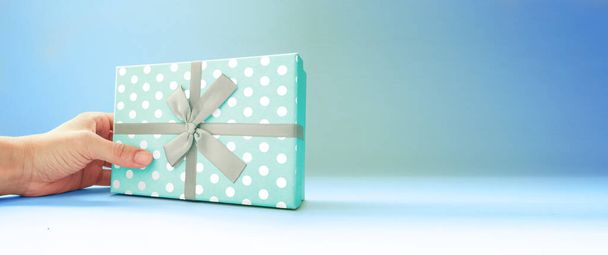 Mano humana sosteniendo la caja de regalo azul, papel de regalo de punto blanco, sobre fondo azul profundo, espacio de copia, vista lateral, para el día del padre, día de la madre, Navidad, cumpleaños. - Foto, imagen