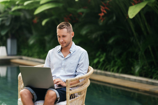 Young man using a laptop computer in a garden with a swimming pool. - Photo, image
