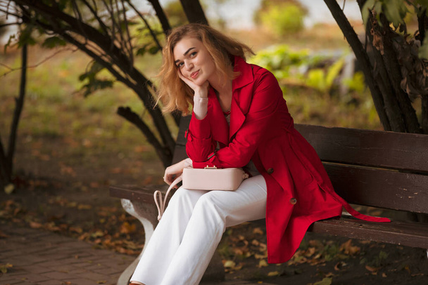 Coquettish woman in red coat sitting on a bench in autumn park leaning head on hand - Foto, imagen