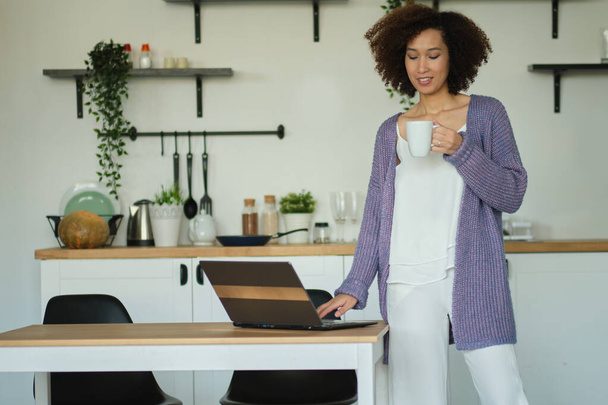 Gelukkige vrouw met een kop koffie en cheques op zijn laptop in haar keuken. Jonge Afro-Amerikaanse vrouw die koffie drinkt in haar keuken. ontspannen ochtend concept - Foto, afbeelding