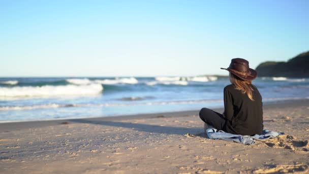 Chłopiec w ciemnych ubraniach siedzi na piaszczystej plaży i patrzy na fale oceanu.. - Materiał filmowy, wideo