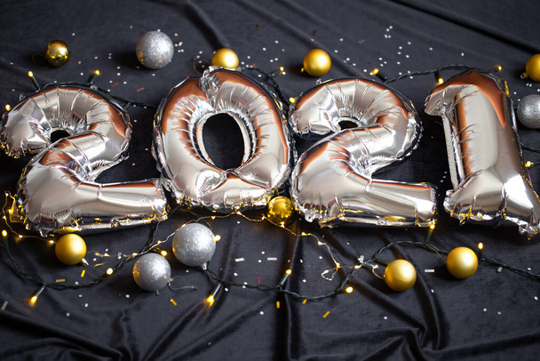 Silver foil balloons made numbers 2021 on black background with garland and balls. Holiday party decoration. Happy new year celebration, congratulation concept. - Fotó, kép