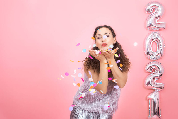 Joyful elegant brunette girl with curly hair in festive clothes throws confetti on a pink background with silver balloons on the right in the form of numbers 2021, new year celebration concept - Photo, Image