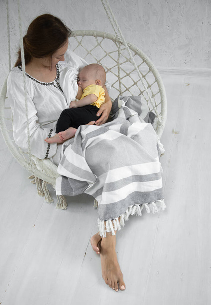 Gelukkig moeder voedt een baby in een witte hangmat met een deken op de achtergrond van een witte kamer. Sluiten.. - Foto, afbeelding