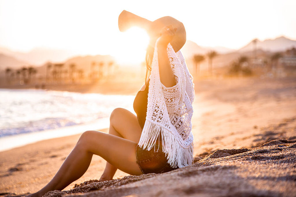 Gyönyörű boho modell fürdőruhában fehér köpennyel a fején pózol a parton naplementekor. - Fotó, kép