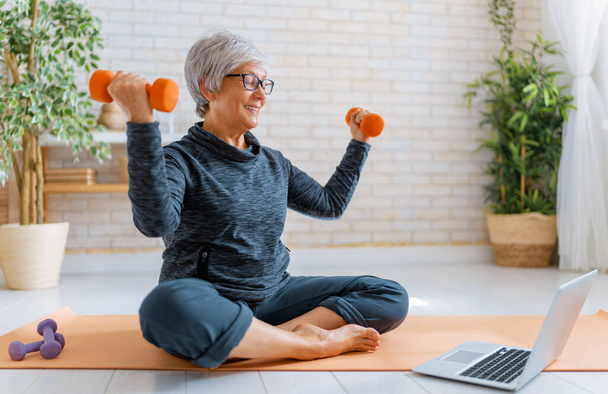 Mujer mayor en ropa deportiva viendo cursos en línea en el ordenador portátil mientras hace ejercicio en casa. - Foto, Imagen