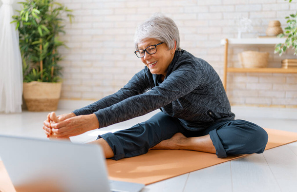 Seniorin in Aktivkleidung sieht sich Online-Kurse am Laptop an, während sie zu Hause trainiert. - Foto, Bild