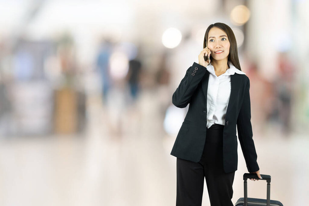 Lächelnde elegante asiatische Geschäftsfrau, die mit ihrem Gepäck auf dem Flughafen unterwegs ist und mit dem Smartphone spricht. - Foto, Bild