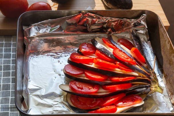 Aubergine crue partiellement tranchée et farcie de tomates et de fromage sur la plaque du four recouverte de papier d'aluminium avant la cuisson, fragment sous un rétroéclairage ensoleillé - Photo, image