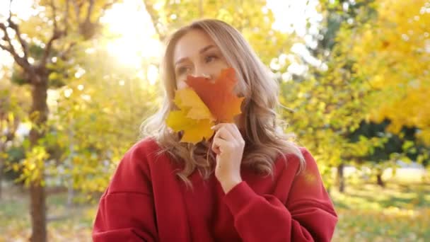 Vrouw in rode jurk verbergt gezicht met gele bladeren in park - Video
