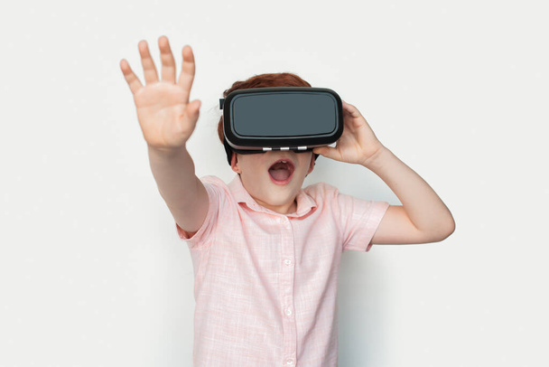 Piccolo ragazzo zenzero caucasico indossa un auricolare realtà virtuale sta toccando qualcosa su un muro di studio bianco - Foto, immagini