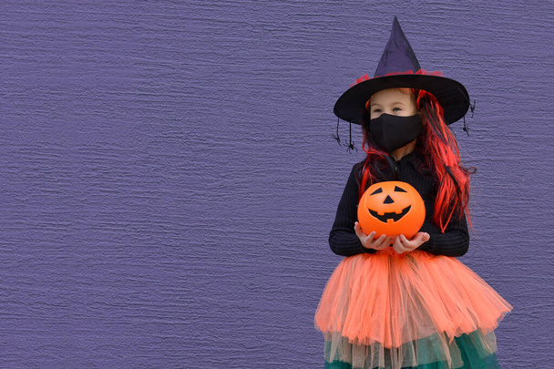 A little girl in a witch costume in a medical mask holding a bucket of pumpkin for sweets on Halloween day. Halloween with safety measures from Covid-19.Copy space for text - Photo, Image