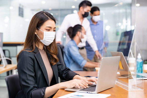 Asyalı ofis çalışanı iş kadını portresi yeni normal ofiste koruyucu yüz maskesi takıyor ve sosyal uzaklık uygulaması Coronavirus COVID-19 'u engellerken ırklar arası takım arka planda yer alıyor.. - Fotoğraf, Görsel