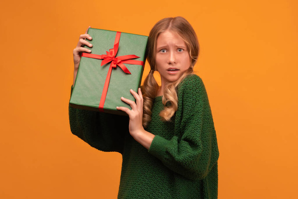 Kép bájos szőke lány 12-14 éves meleg zöld pulóverben tartja jelen doboz piros íj. Stúdiófelvétel, sárga háttér, elszigetelt. Újév Női Nap születésnapi ünnep koncepció - Fotó, kép