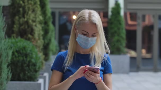 Portrait of a charming young woman in a protective medical mask is texting message and talking on her smartphone. Corona virus health protection concept. Slow motion. Close up. - Imágenes, Vídeo