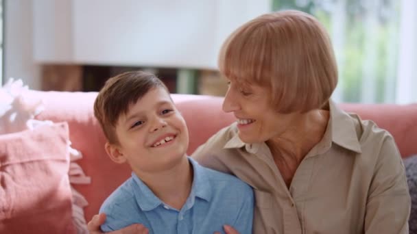 Grootmoeder knuffelt kleinzoon in de kamer. Glimlachende jongen aan het praten met oudere vrouw - Video