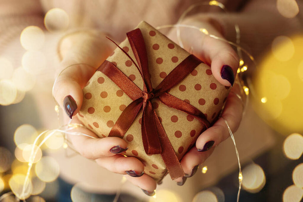 Hände halten und verschenken aus gebastelter brauner Verpackung. Ein Mädchen in einem rosafarbenen Strickpullover hält ein Neujahrs- und Weihnachtsgeschenk mit einer Girlande mit gelben Lichtern in der Hand - Foto, Bild