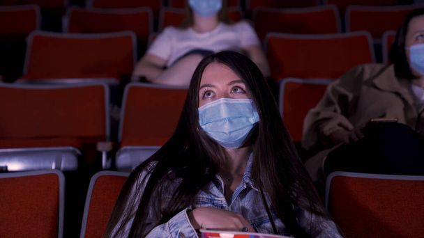 Elokuva karanteenissa, sepelvaltimoviruspandemian säännöt, sosiaalinen etäisyys elokuvien katselun aikana. Media. Kaunis pitkäkarvainen nuoria naisia suojaava kasvonaamio istuu rivissä auditorio - Valokuva, kuva