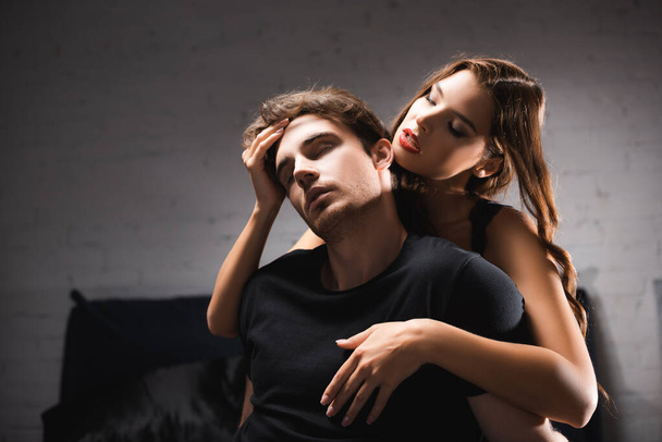 Sexy woman with red lips hugging and touching young man with closed eyes at home  - Zdjęcie, obraz